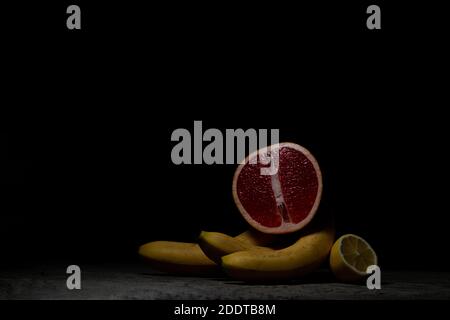 A juicy red grapefruit rests on three bananas, next to a lemon on gray concrete. Low key. Stock Photo