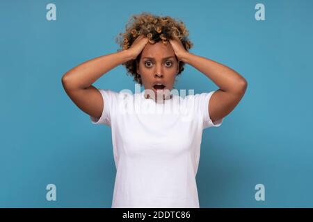 Surprised young african woman holding her head in hands in full disbelief while standing isolated on blue wall. Stock Photo