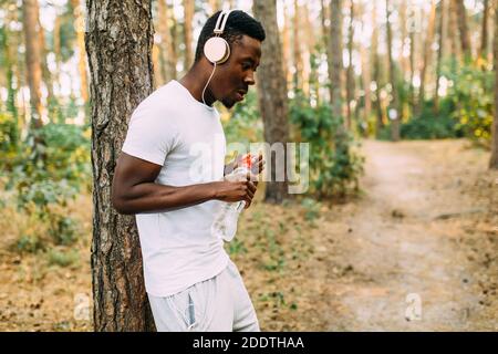 A young athletic African American relaxes after an intense workout. A man rests with a bottle of water, leaning against a tree, after a morning jog Stock Photo