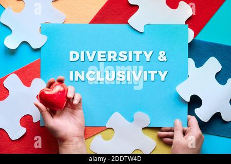 Hand with red stone heart. Text Diversity and Inclusivity on paper. Felt, white puzzle pieces. Stock Photo