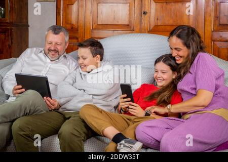 Happy family sitting on sofa and using digital tablet and mobile phone at home - Father and son laughing while watching tha tablet and mother and daug Stock Photo