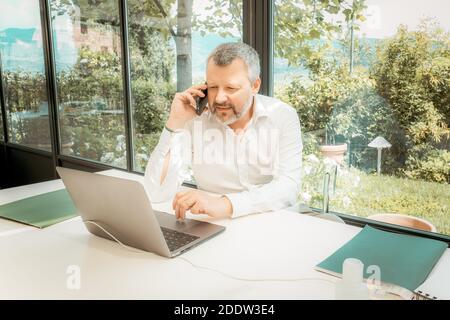 Man is talking on his cell phone and using laptop, working at home. Concerned about finances. Handsome man with beard working at home Stock Photo