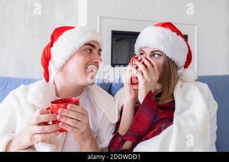 Christmas at home concept. Profile side view photo of two lovely, carefree persons sitting on cozy couch or sofa in living room holding red cups looki Stock Photo