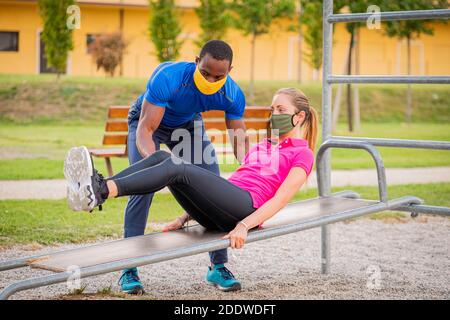 Afro american fitness instructor coaching helping woman doing push-ups on cross fit horizontal bar in the park wearing medical protective masks - Youn Stock Photo