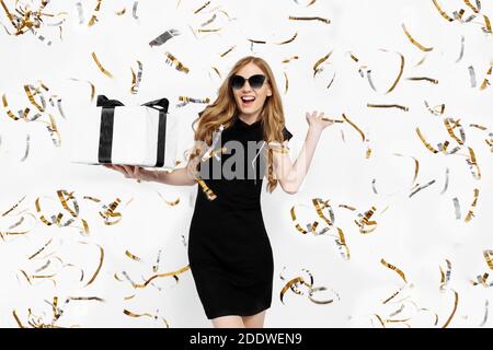 Happy excited young girl in black dress and sunglasses holding gift box with black ribbon having fun on white background with festive confetti. Shoppi Stock Photo