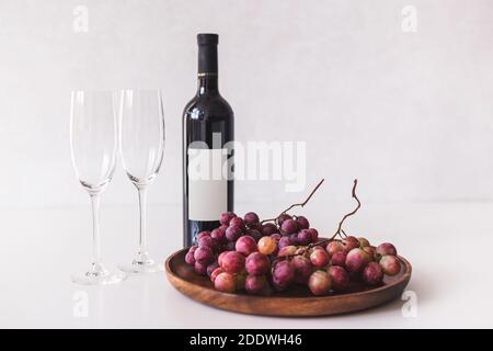 Red wine bottle, large burgundy fresh grapes on wooden dish, empty wine glasses on white background, mockup