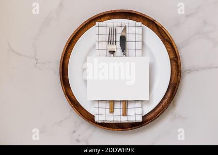 Blank paper greeting card on wooden dish, cutlery with checkered napkin, cotton flowers. White marble background. Flat lay, top view Stock Photo