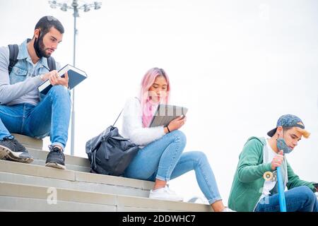 Young people university studyng outoor during coronavirus pandemic. Four multietnic university students with protective masks sitting outdoor Stock Photo