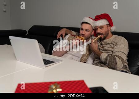 Young caucasian gay couple with Santa Claus hats watching movies on the laptop and eating pizza on the Christmas Eve. High quality photo Stock Photo