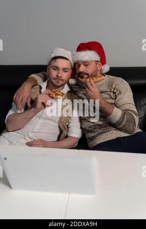 Handsome homosexual couple with Santa Claus hats watching movies and eating pizza on Christmas eve. High quality photo Stock Photo