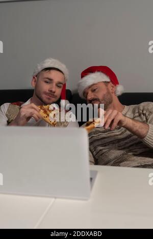 Young caucasian gay couple with Santa Claus hats watching movies on the laptop and eating pizza on the Christmas Eve. High quality photo Stock Photo