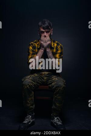Picture on a black background of a bearded male model sitting on a chair and covering his face with his tattooed hands Stock Photo