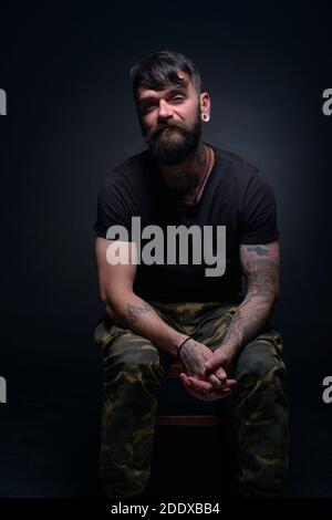 Brutal attractive bearded man with tattooed hands poses in black blank t-shirt and army pants seated on a chair Stock Photo