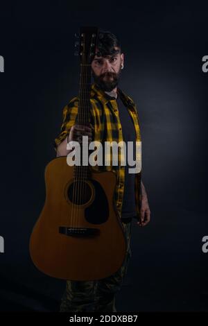 Tattooed man with a beard wearing a tartan yellow black shirt is standing and posing with his guitar in his hand Stock Photo