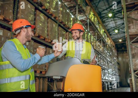 Warehouse workers in orange helmets standing near shelves and having coffee Stock Photo