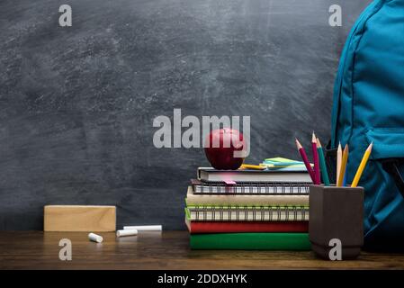 Stack of books, colorful stationery and education supplies on wooden table in classroom with blackboard in background Stock Photo