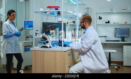 Scientist working in modern equipped laboratory cheching virus development with microscope. Multiethnic team examining vaccine evolution using high tech and chemistry tools for scientific research. Stock Photo