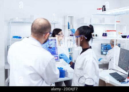Scientist holding glass flask with blue genetic material in busy laboratory. Multiethnic team of medical researchers working together in sterile lab wearing protection glasses and gloves. Stock Photo
