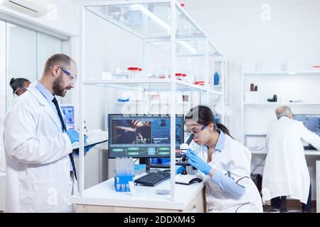 Male scientist taking notes on tablet pc during virus study. Team of researchers doing pharmacology engineering in sterile laboratory for healthcare industry with african assistant in the background. Stock Photo