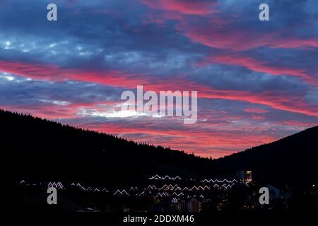 Scenic dramatic fiery cloudy sunset evening view over mountain resort village Bukovel in Ukraine with roof lights and forest tree silhouettes Stock Photo