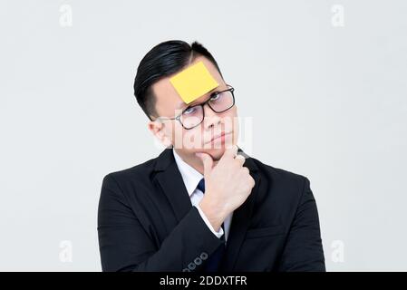 Bored and curious  Asian businessman thinking with sticky note paper on forehead isolated on light gray background Stock Photo