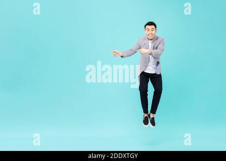 Young smiling Asian man with funny face jumping and pointing hands to copy space aside on studio light blue background Stock Photo