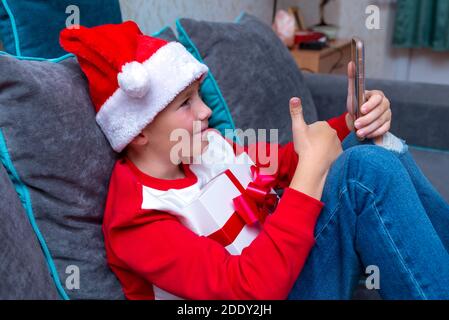 Handsome kid boy in Santa Claus hat looks at smartphone screen, shows thumb up.Talk with family,closeup image. Stock Photo