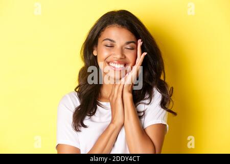 Close-up of dreamy, beautiful african-american woman, gently touching face and smiling pleased, standing against yellow background Stock Photo