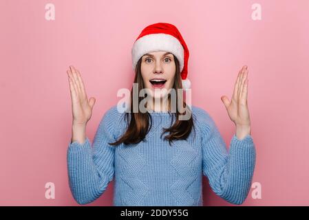 Shocked cute woman in Christmas hat shows something large and of big size, measures with hands and keeps mouth opened from wonder Stock Photo