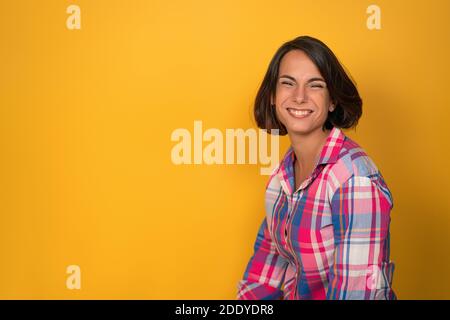 Super excited young woman with brown hair and plaid long sleeve shirt extremely smiling on camera. Isolated on yellow background Stock Photo