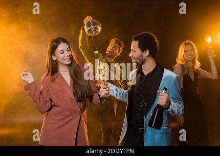 happy multicultural couple holding glasses of champagne and bottle near cheerful friends on blurred and black background Stock Photo