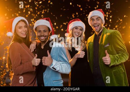 smiling interracial friends in santa hats showing thumbs up near falling confetti on black Stock Photo