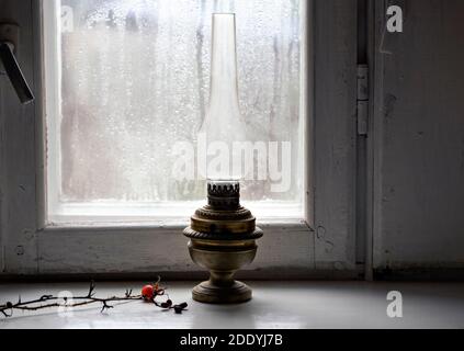 Soft focus on vintage oil lamp on the windowsill with twig in blurred rainy window background. Close up Stock Photo
