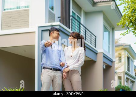Portrait of Asian young couple standing and hugging together and holding house key looking happy in front of their new house to start new life. Family Stock Photo