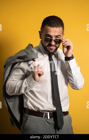 Bearded and serious business person dressed in greyish suite posing holding his jacket on his shoulder hanging it behind looking lowering sunglasses Stock Photo