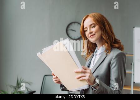 Smiling businesswoman looking at folder with paper sheets with blurred office on background Stock Photo