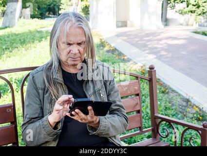 elderly man opens application on smartphone while sitting on bench in city park on sunny autumn day Stock Photo