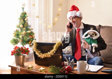 Man in a suit with a Christmas hat working in the living room at Christmas holidays with his dog in his arms. Front view. Horizontal composition. Stock Photo