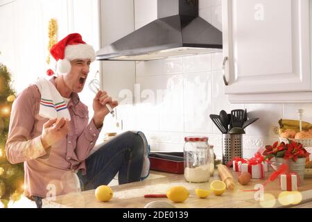 Funny man cooking with christmas hat in kitchen with pastry ingredients on bench and decorated tree in the background. Horizontal composition. Stock Photo