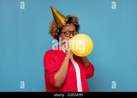Astonished african woman blowing air baloon for birthday party. Studio shot on blue wall. Stock Photo