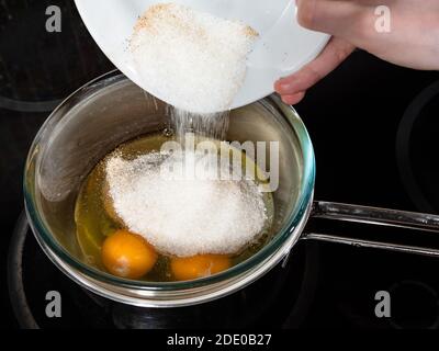cooking sweet sponge cake at home - adding sugar with cinnamon to raw eggs in glass bowl on water bath on stove at home kitchen Stock Photo