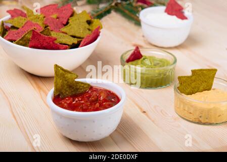Christmas festive snacks for the party. Green and Red Tortilla chips in the shape of a Christmas tree in bowls with salsa, guacamole, cheese, and mayo Stock Photo