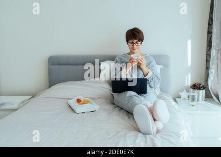 Caucasian woman in glasses, casual home clothes drinking her breakfast coffee during working on laptop on her bed. Remote working from home. Time at Stock Photo