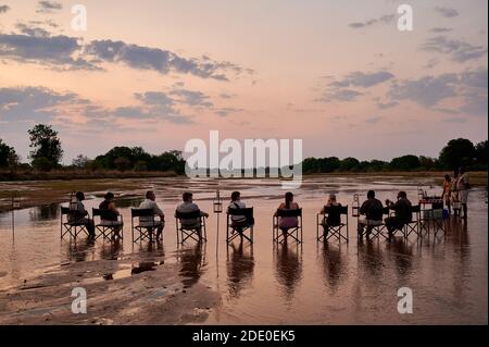 Tourists sittting on chairs in the Kapamba River while taking a sundowner in the sunset, South Luangwa National Park, Mfuwe, Zambia, Africa Stock Photo