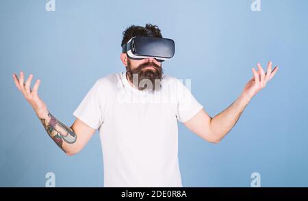 Man with hipster beard in idk posture isolated on blue background. Bearded software developer clueless about program bug in new VR equipment. Bearded man in virtual reality glasses testing new gadget. Stock Photo