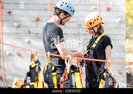 Male instructor puts protective climbing equipment on a young woman at amusement park outdoors Stock Photo