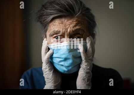 Portrait senior woman holding head hands, sad worried emotion in protective medical mask, gloves against virus and infection at home. Old gray hair fe Stock Photo