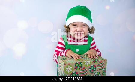 Little elf with a cute smile is sitting on the white background and playing or packing Christmas presents. Christmas holiday background photo. Stock Photo