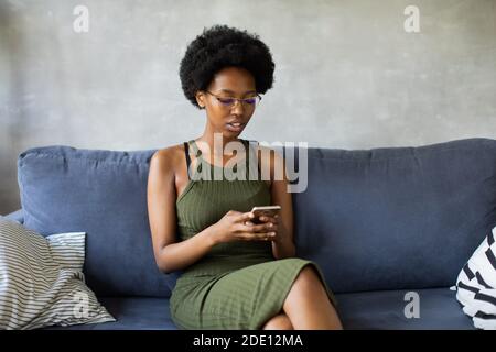 Happy african American young woman sit relax on cozy couch and writing on the phone. Happy to move to new apartment. Stock Photo