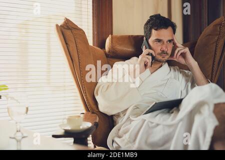 A man in a white bathrobe is having a call  from the hotel room. Seating in a comfortable lather armchair with a tablet on his knees and coffee Stock Photo